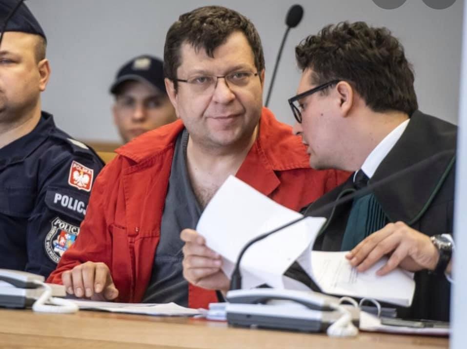 Mężczyzna w czerwonej kurtce, prawdopodobnie oskarżony, siedzi obok adwokata w todze w sali sądowej, trzymając dokumenty. W tle widoczni policjanci.