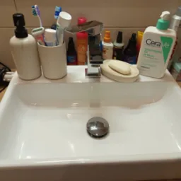 Umywalka z licznymi kosmetykami i akcesoriami higienicznymi, w tym mydło, żel do mycia twarzy CeraVe, szczoteczki do zębów i suszarka do włosów. Ujęcie z bliska.