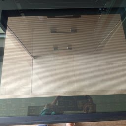 Clean For You usługi sprzątające Paulina Tarasiuk - Czyszczenie piekarnika z widoczną szybą i odbiciem dłoni trzymającej ściereczkę. Widoczne ustawienia temperatury i funkcji na szkle piekarnika.