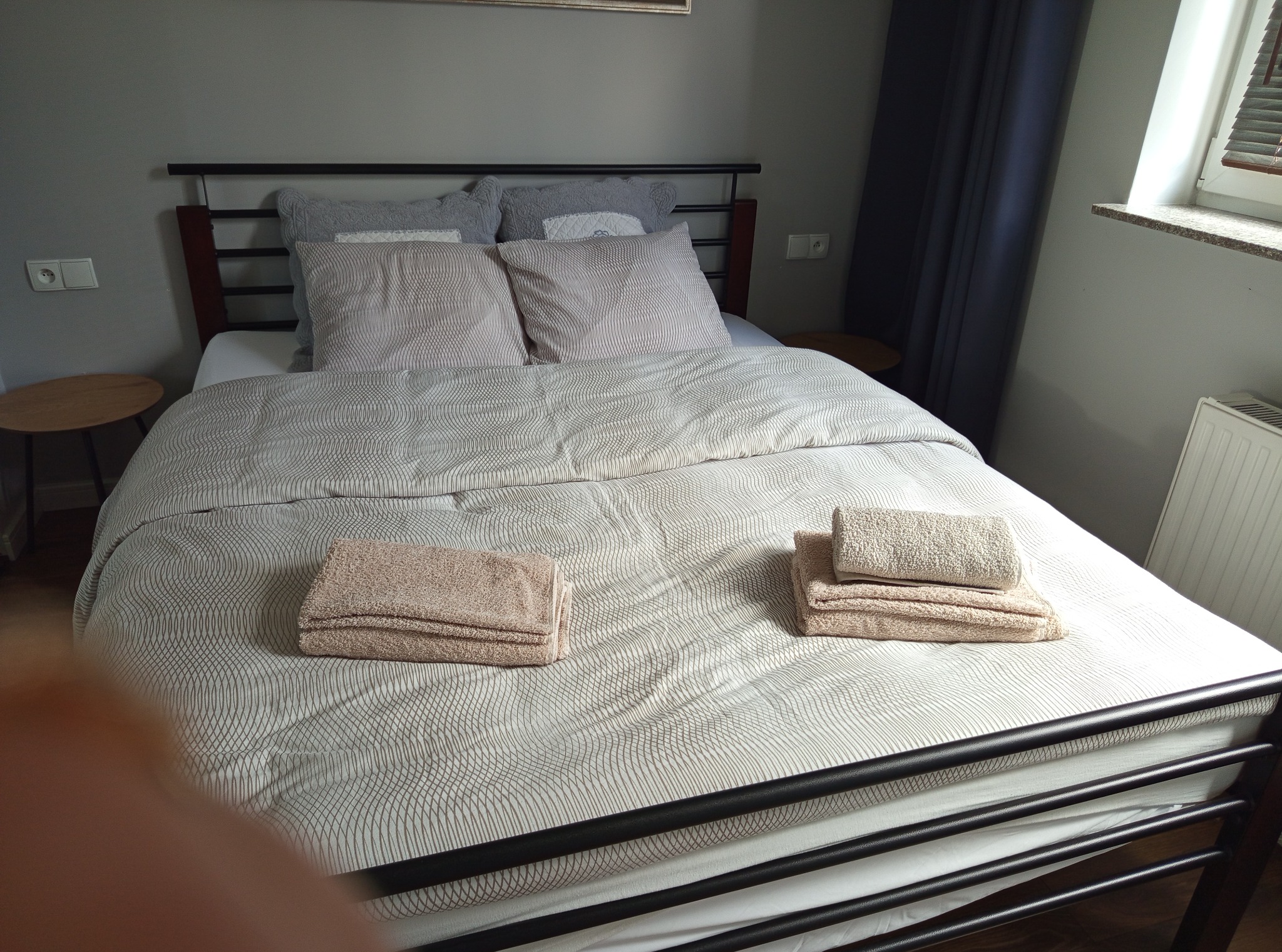 Czyste, poskładane ręczniki na świeżo pościelonym łóżku z metalową ramą w pokoju z oknem i grzejnikiem. Widok z góry, neutralne kolory.