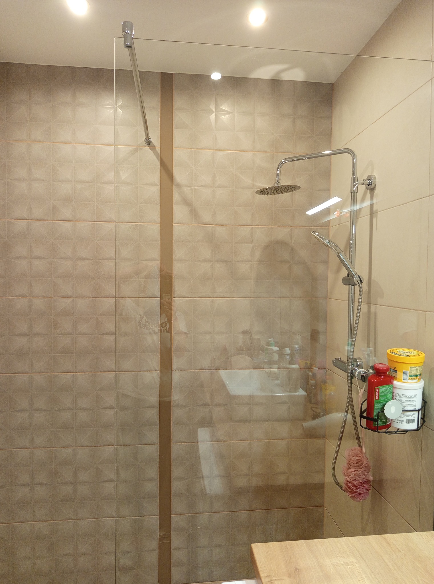 Beżowa kabina prysznicowa z deszczownicą i półką na kosmetyki. Widoczne płytki z delikatnym wzorem i szklana ściana kabiny. Oświetlenie sufitowe.