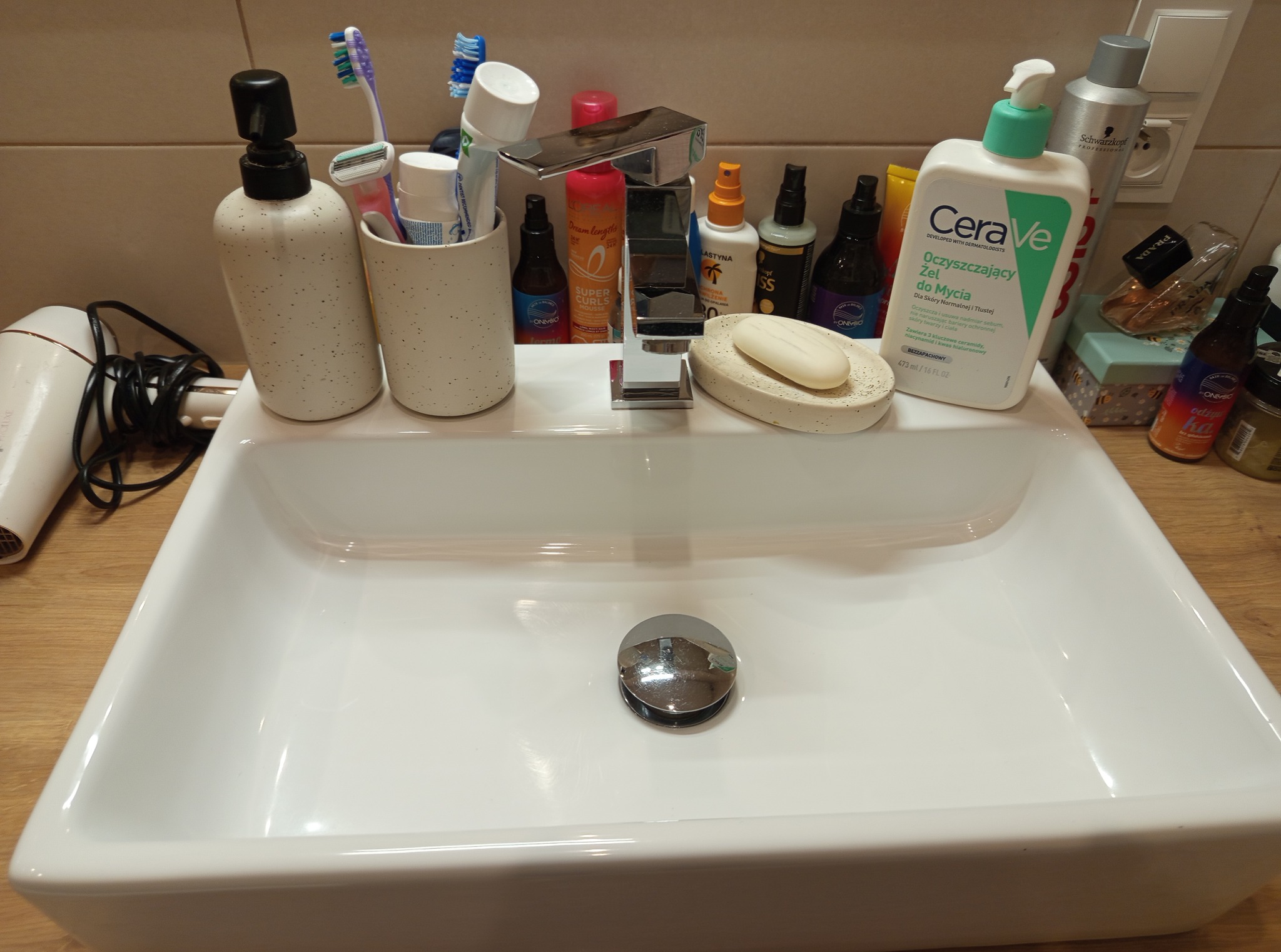 Umywalka z licznymi kosmetykami i akcesoriami higienicznymi, w tym mydło, żel do mycia twarzy CeraVe, szczoteczki do zębów i suszarka do włosów. Ujęcie z bliska.