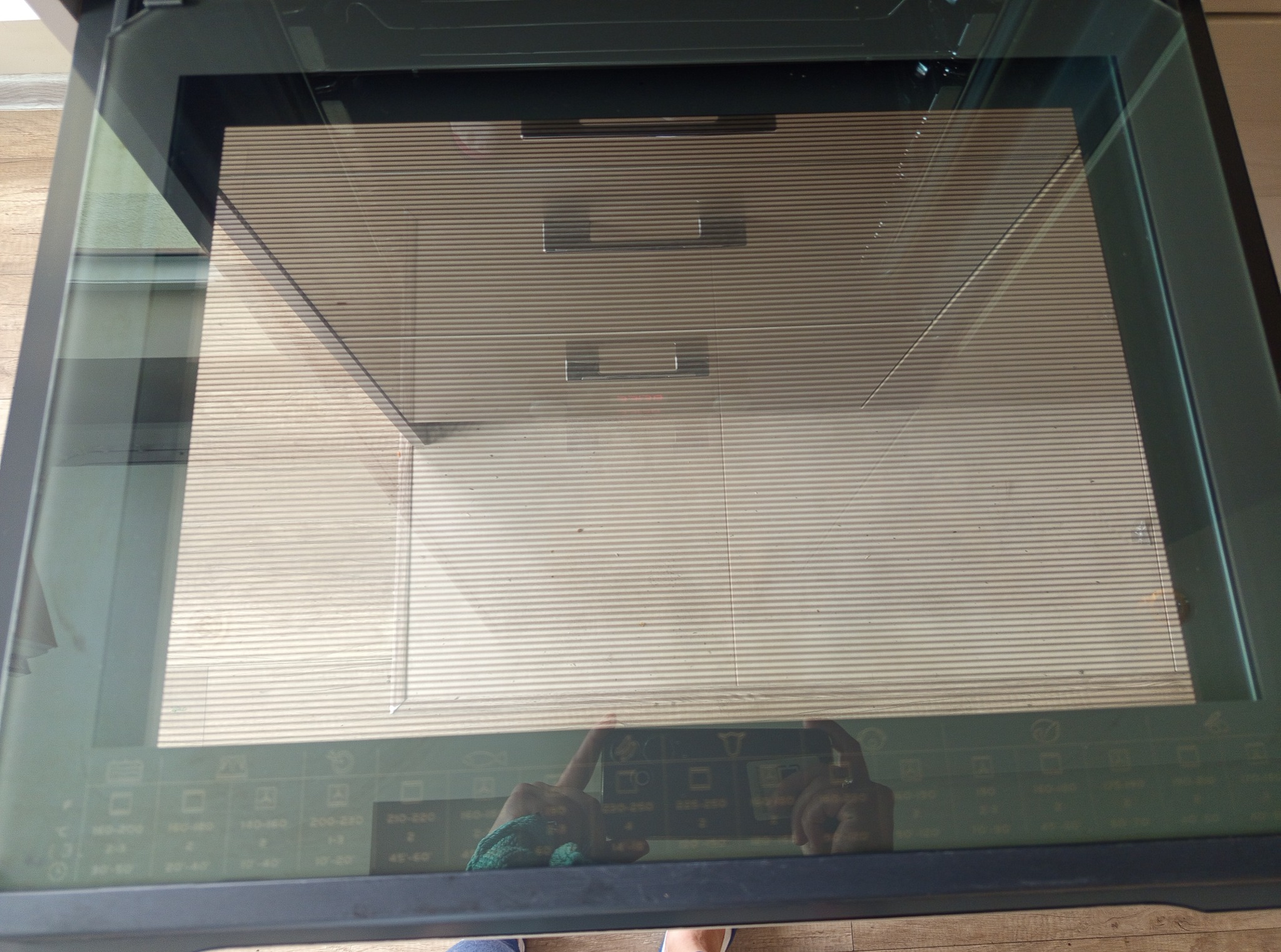 Czyszczenie piekarnika z widoczną szybą i odbiciem dłoni trzymającej ściereczkę. Widoczne ustawienia temperatury i funkcji na szkle piekarnika.