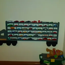 Półka na kolekcję autek w kształcie ciężarówki, szara, zamontowana na białej ścianie. Na półkach ustawione modele aut, na dole zabawka pluszowa leżąca na wózku.