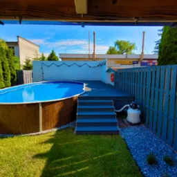 Owalny basen naziemny z drewnopodobną obudową, otoczony zielenią i szarą drewnianą konstrukcją z drabinką i małym domkiem ratownika z kołem ratunkowym.