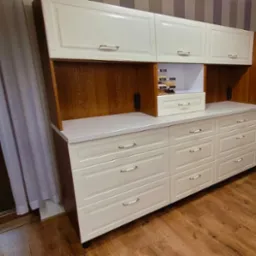 Biała, klasyczna zabudowa kuchenna z drewnianymi akcentami, szufladami i szafkami. Widoczna półka z przyprawami i sprzęt AGD. Ujęcie z perspektywy podłogi.