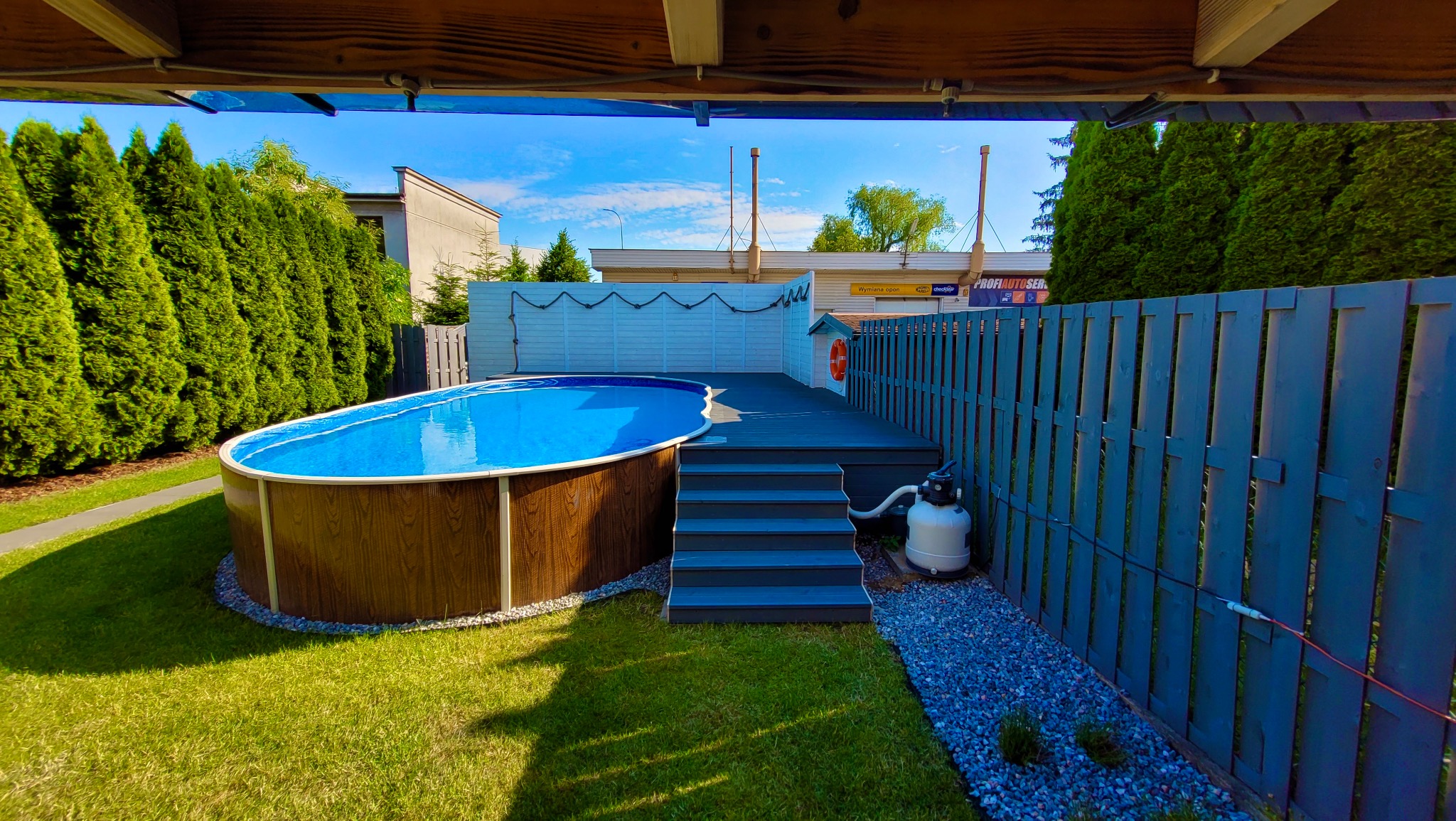 Owalny basen naziemny z drewnopodobną obudową, otoczony zielenią i szarą drewnianą konstrukcją z drabinką i małym domkiem ratownika z kołem ratunkowym.