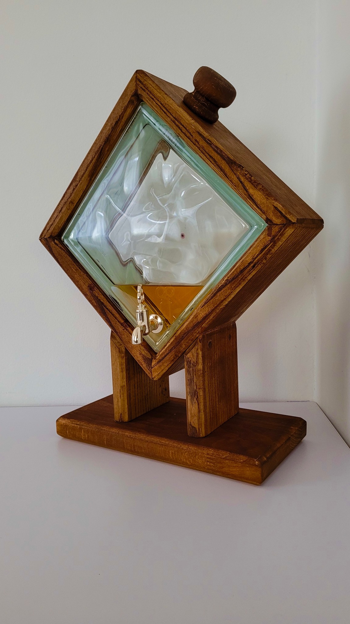 Karafka w drewnianej, kwadratowej ramie, ustawiona pod kątem. Widoczny kranik i podstawa z drewna. Wewnątrz płyn o bursztynowej barwie. Całość na białym blacie.