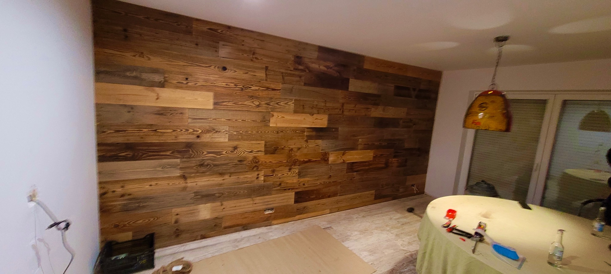 Ściana wykończona panelami z postarzanego drewna, widoczne narzędzia na stole, wnętrze w trakcie prac wykończeniowych, lampa wisząca.