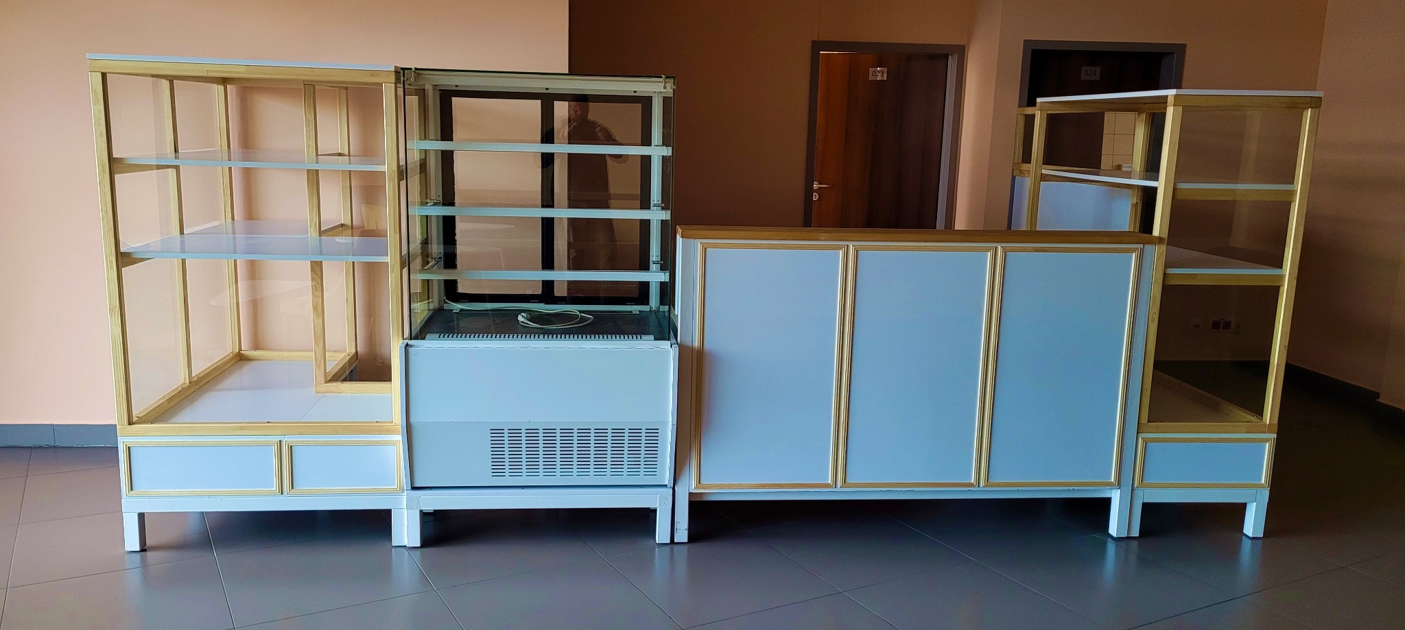 Komplet mebli sklepowych z chłodziarką, drewnianą ramą i szklanymi półkami, ustawiony na szarej podłodze, na tle beżowej ściany z drzwiami.