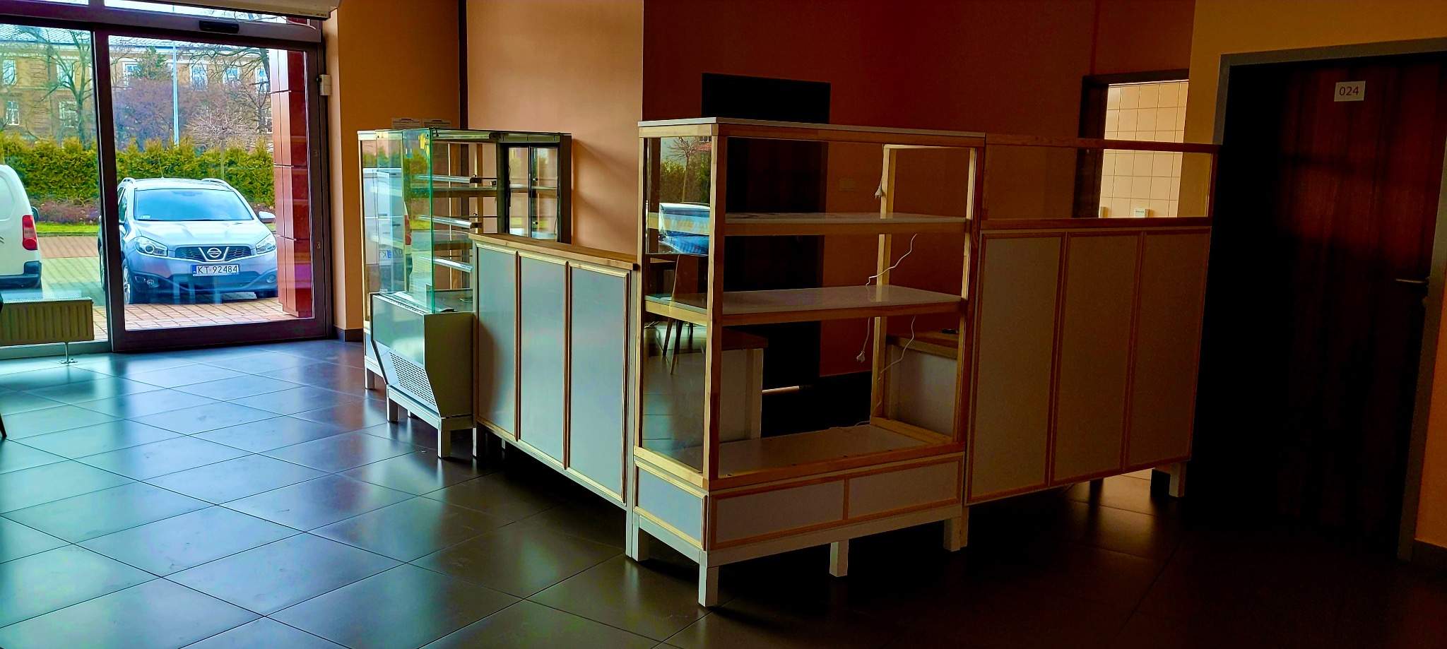 Wnętrze z częściowo zmontowanymi szafami i witrynami, widoczne elementy konstrukcyjne i wykończeniowe, podłoga z płytek, drzwi wejściowe z widokiem na zewnątrz.