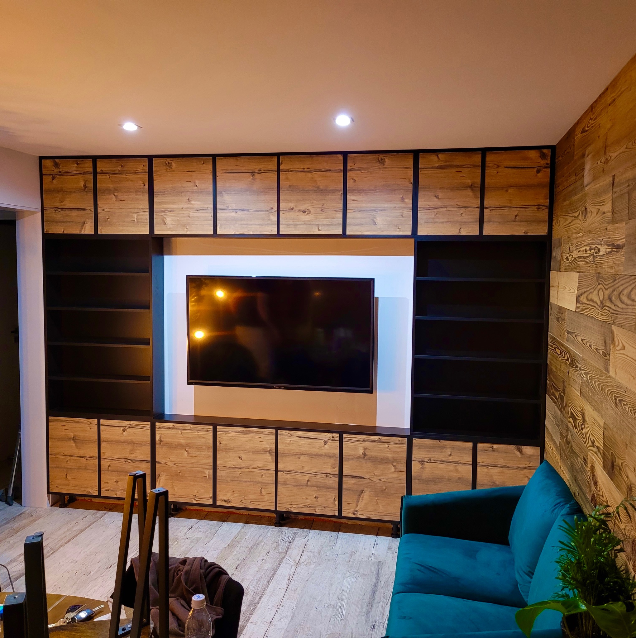 Drewniana szafa na wymiar z czarnymi elementami metalowymi, wnęką na telewizor i ścianą z paneli drewnianych. W tle widoczny fragment sofy.