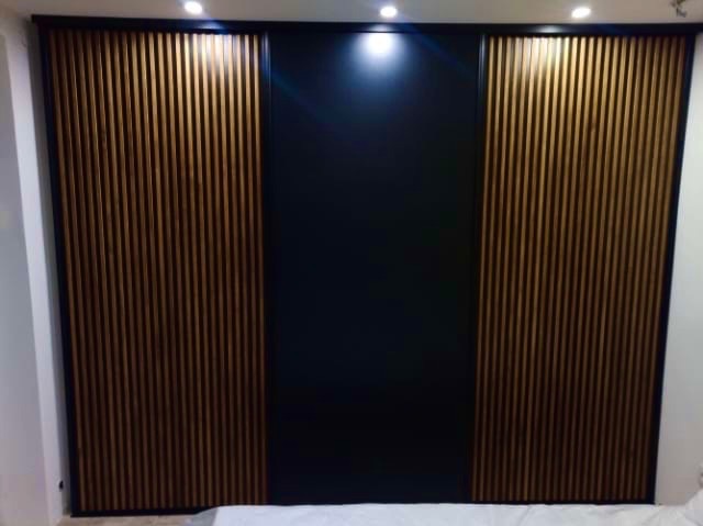 Nowoczesna szafa z przesuwnymi drzwiami: czarny panel centralny, drewniane lamele po bokach, oświetlenie punktowe na górze. Minimalistyczny design.