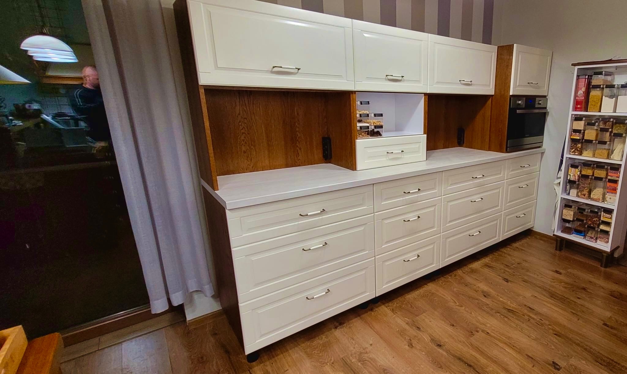 Kremowe szafki kuchenne z drewnianymi akcentami i szufladami, uzupełnione o półki z przyprawami, na tle drewnianej podłogi i zasłoniętego okna.