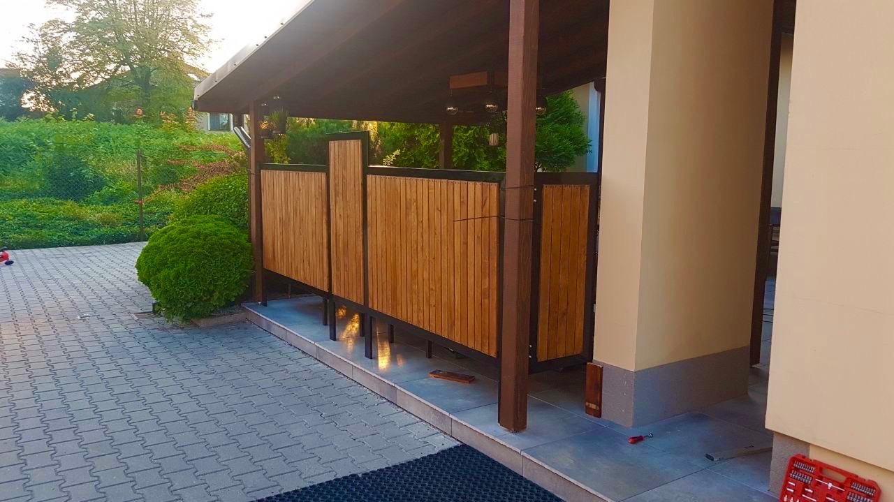 Drewniana pergola z elementami zadaszenia i zabudowy bocznej, zlokalizowana w Tarnowie. Widoczna kostka brukowa i zieleń w tle. Detale konstrukcyjne.