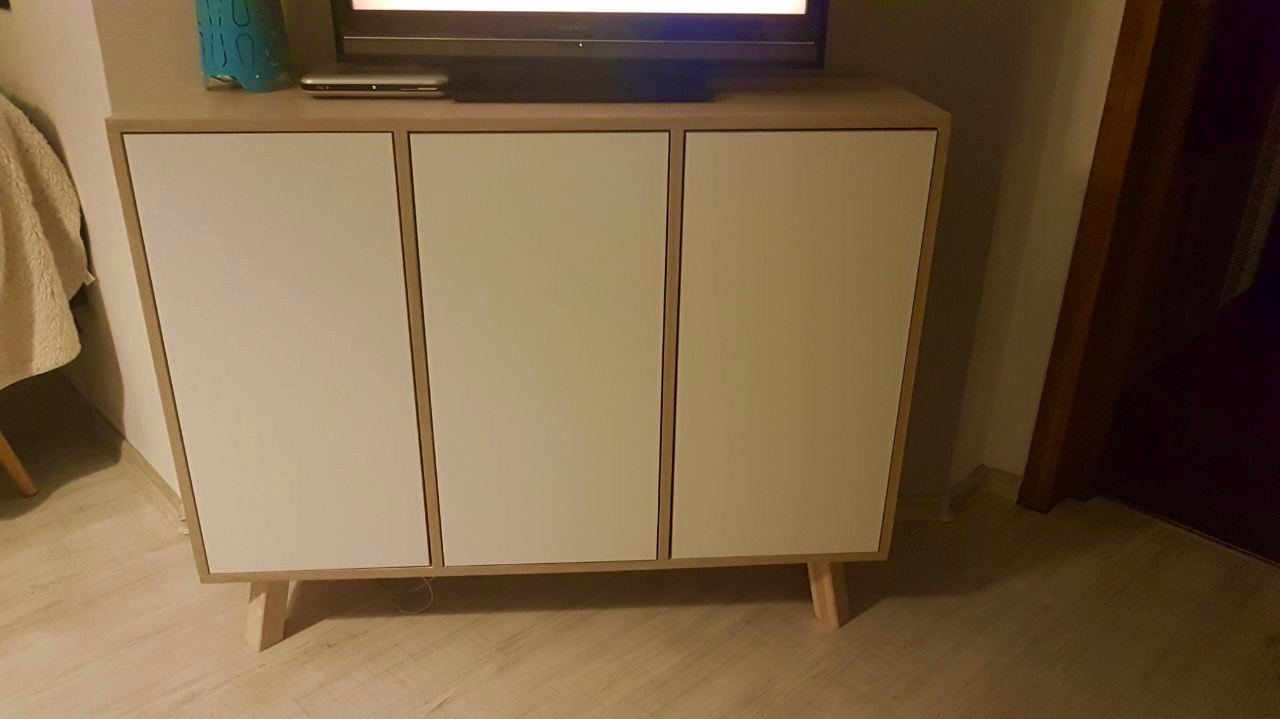 Nowoczesna szafka RTV z trzema białymi frontami i jasną drewnianą ramą, ustawiona na drewnianej podłodze, z telewizorem na blacie.