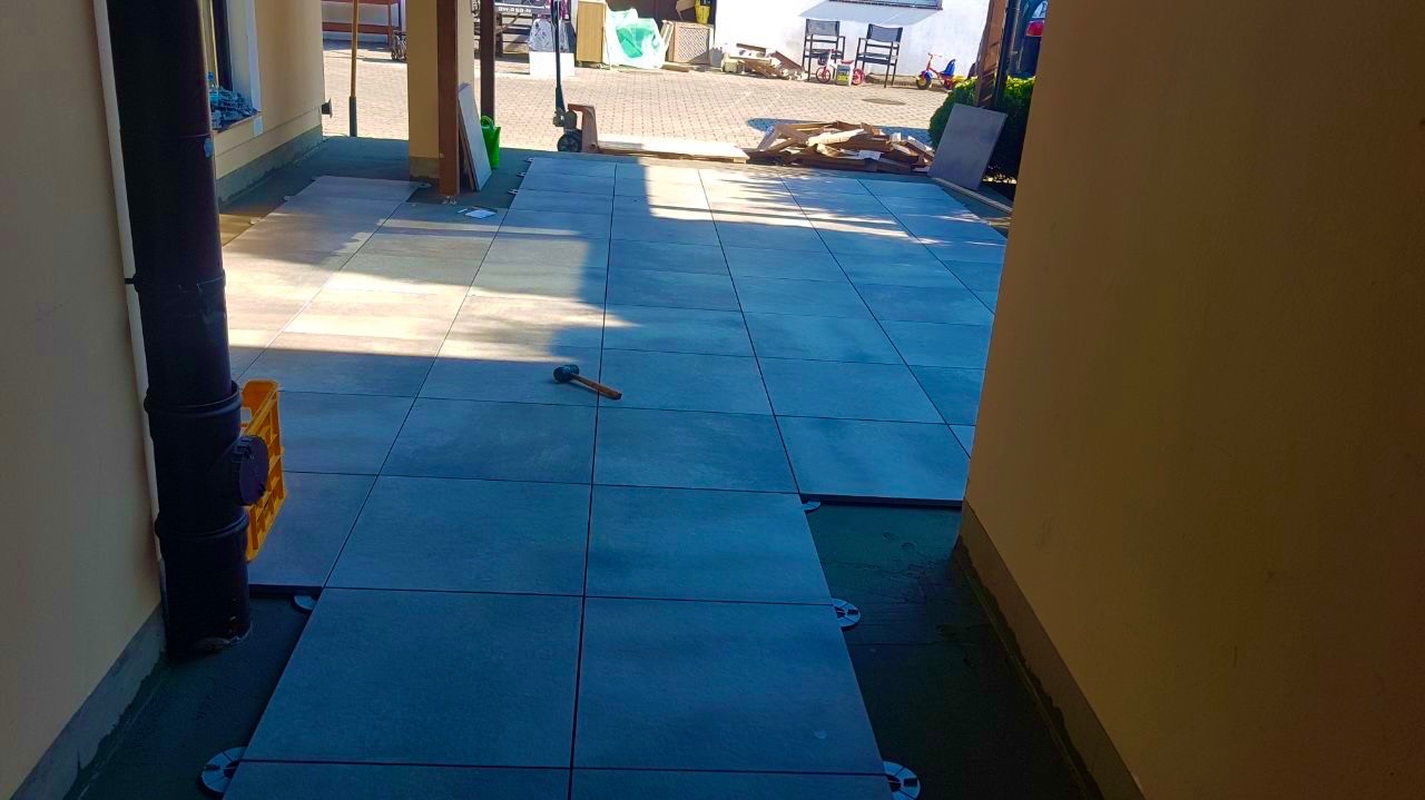 Układanie płytek tarasowych na wspornikach, widoczny młotek i częściowo ułożony taras. Na zewnątrz domu, w tle elementy otoczenia.