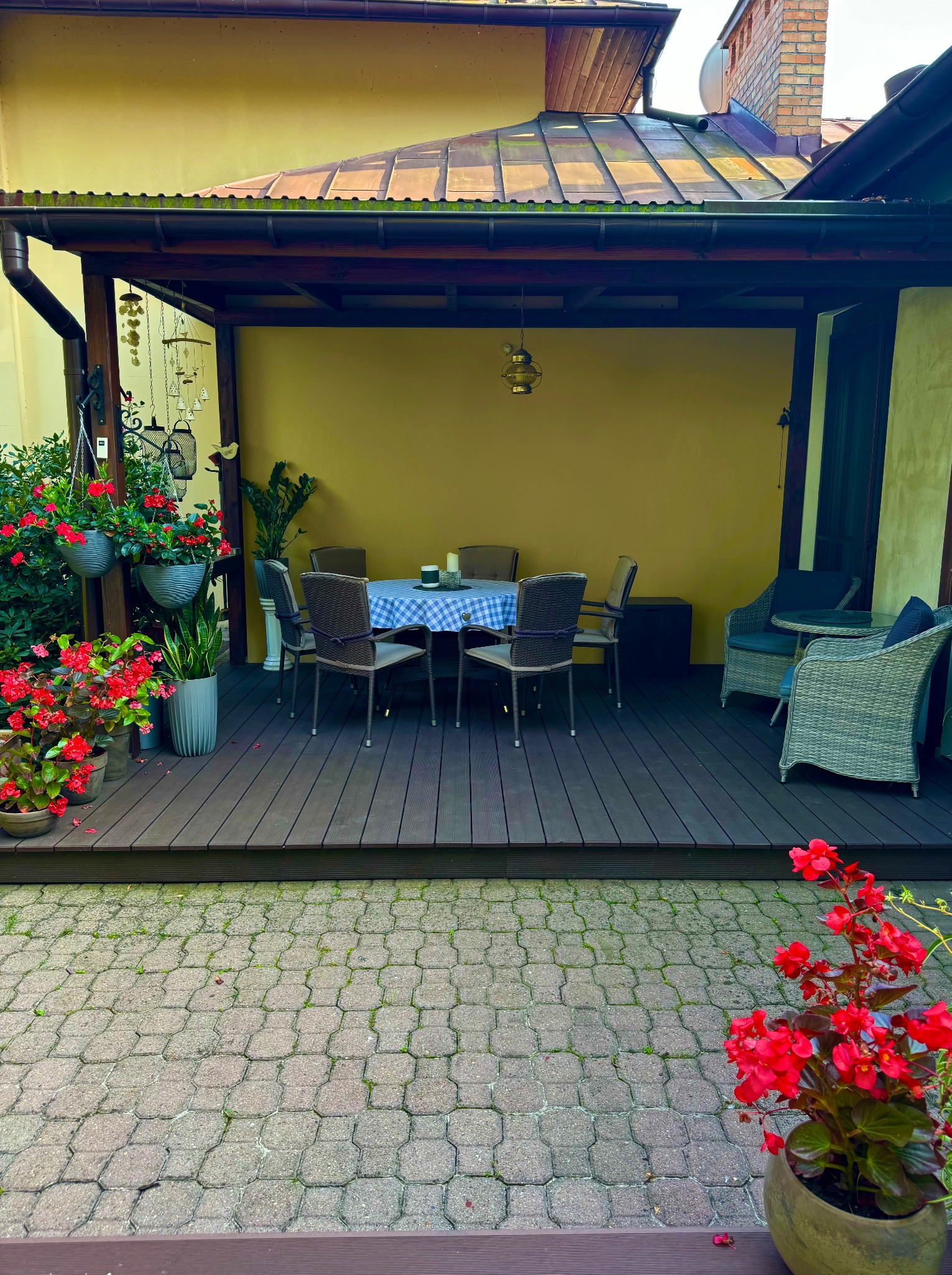 Zadaszony taras z ciemnego drewna. Stół z krzesłami, dekoracje kwiatowe w donicach. W tle żółta ściana i elementy architektoniczne.