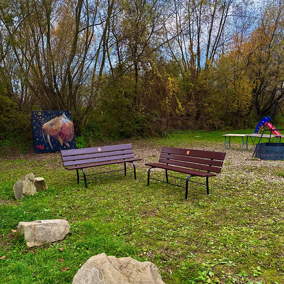 Dwie drewniane ławki parkowe na trawie, obok drzew i placu zabaw. Na pierwszym planie kamienie, w tle obraz z motywem zwierzęcym.
