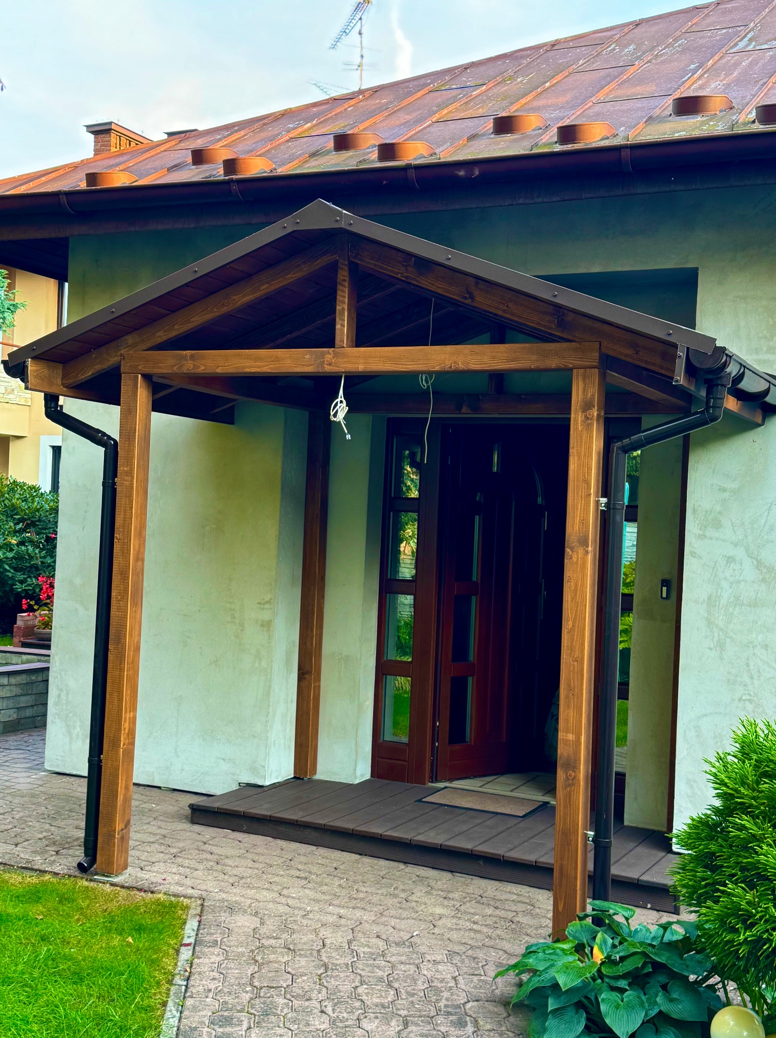 Drewniany daszek nad wejściem do domu, z widocznymi elementami konstrukcyjnymi i detalami wykończenia. Schody wejściowe wyłożone deskami. Elewacja domu w jasnym odcieniu.