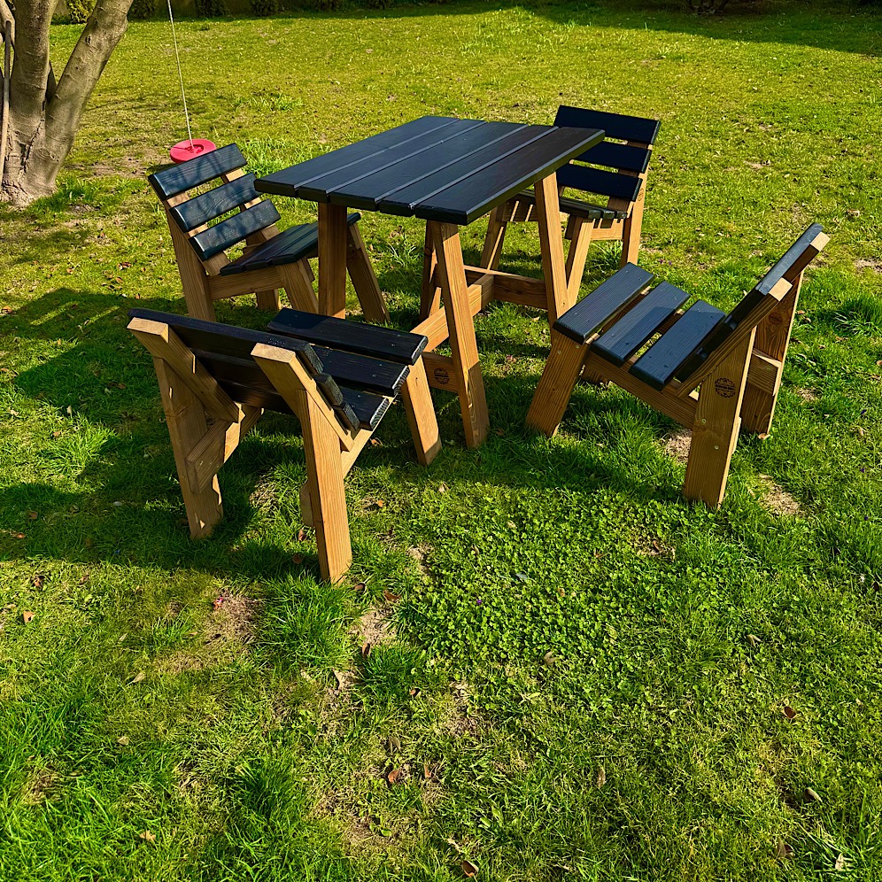 Stół i cztery ławki ogrodowe z drewna, pomalowane na czarno, stoją na trawniku w słoneczny dzień. Widoczne detale konstrukcji i faktura drewna.