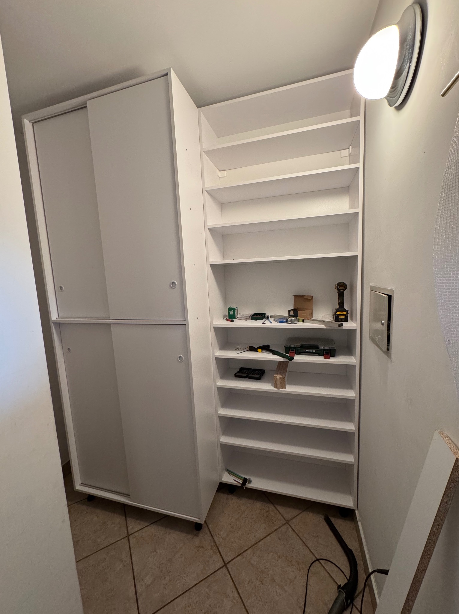 Biała szafa na wymiar z przesuwnymi drzwiami i otwartymi półkami, w trakcie montażu, narzędzia widoczne na półkach, ciepłe światło lampy.