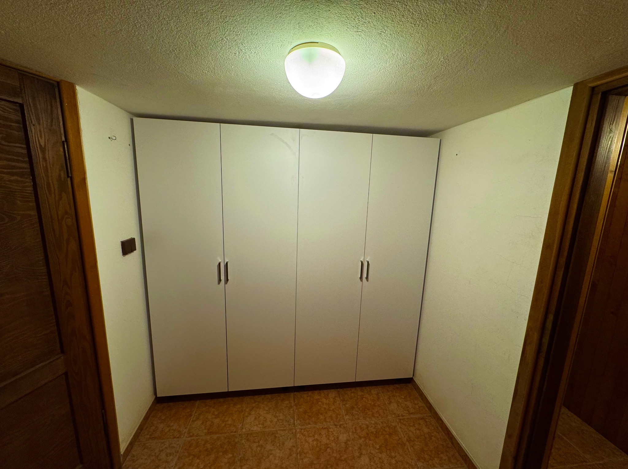 Wnętrze z czterodrzwiową szafą wnękową, białe fronty, minimalistyczne uchwyty, oświetlenie sufitowe, brązowe płytki podłogowe, widok z perspektywy wejścia.