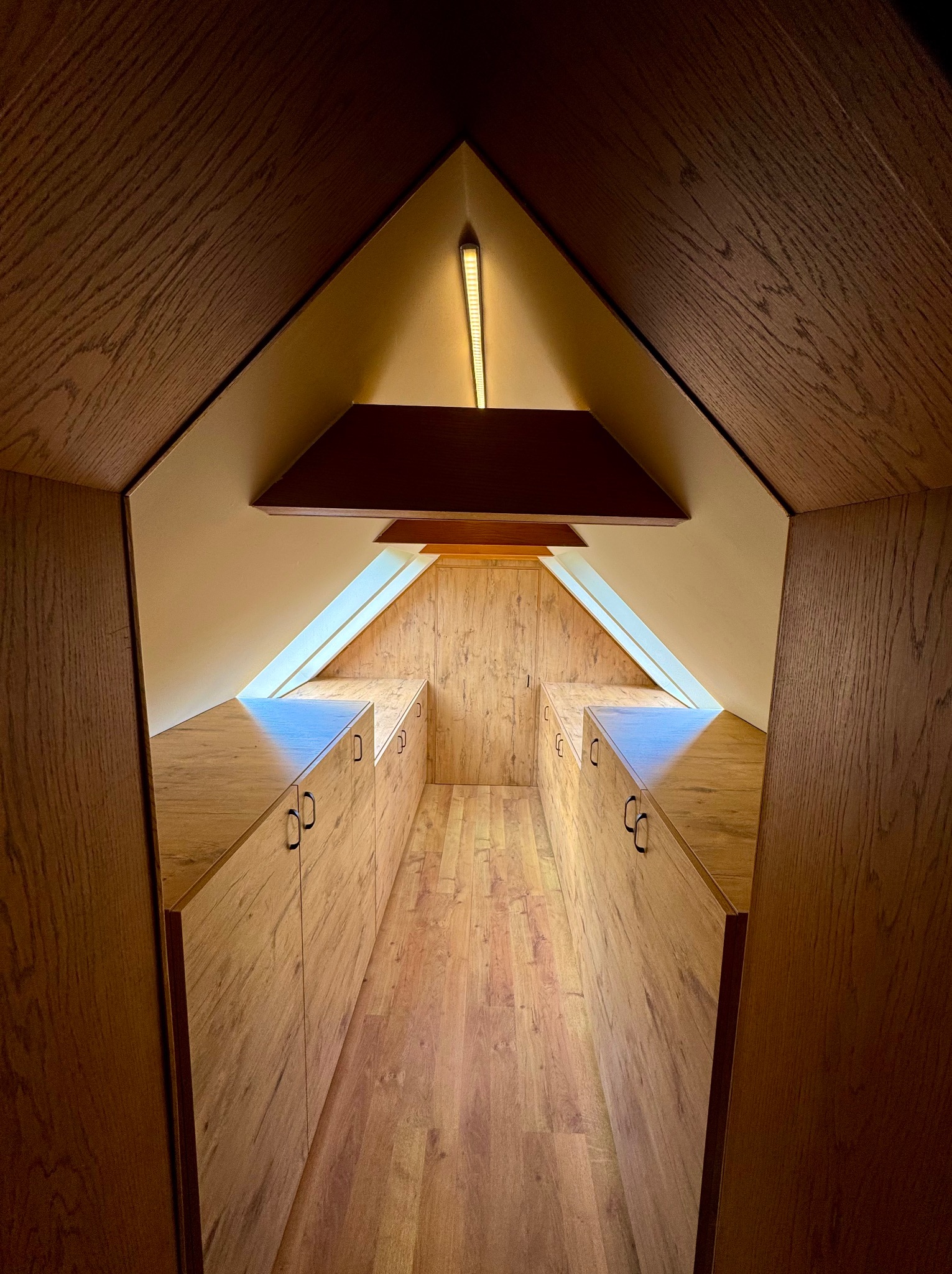 Szafa na wymiar na poddaszu ze skosami, wykończona drewnem w ciepłym odcieniu, z oświetleniem punktowym. Nietypowy układ i zabudowa.