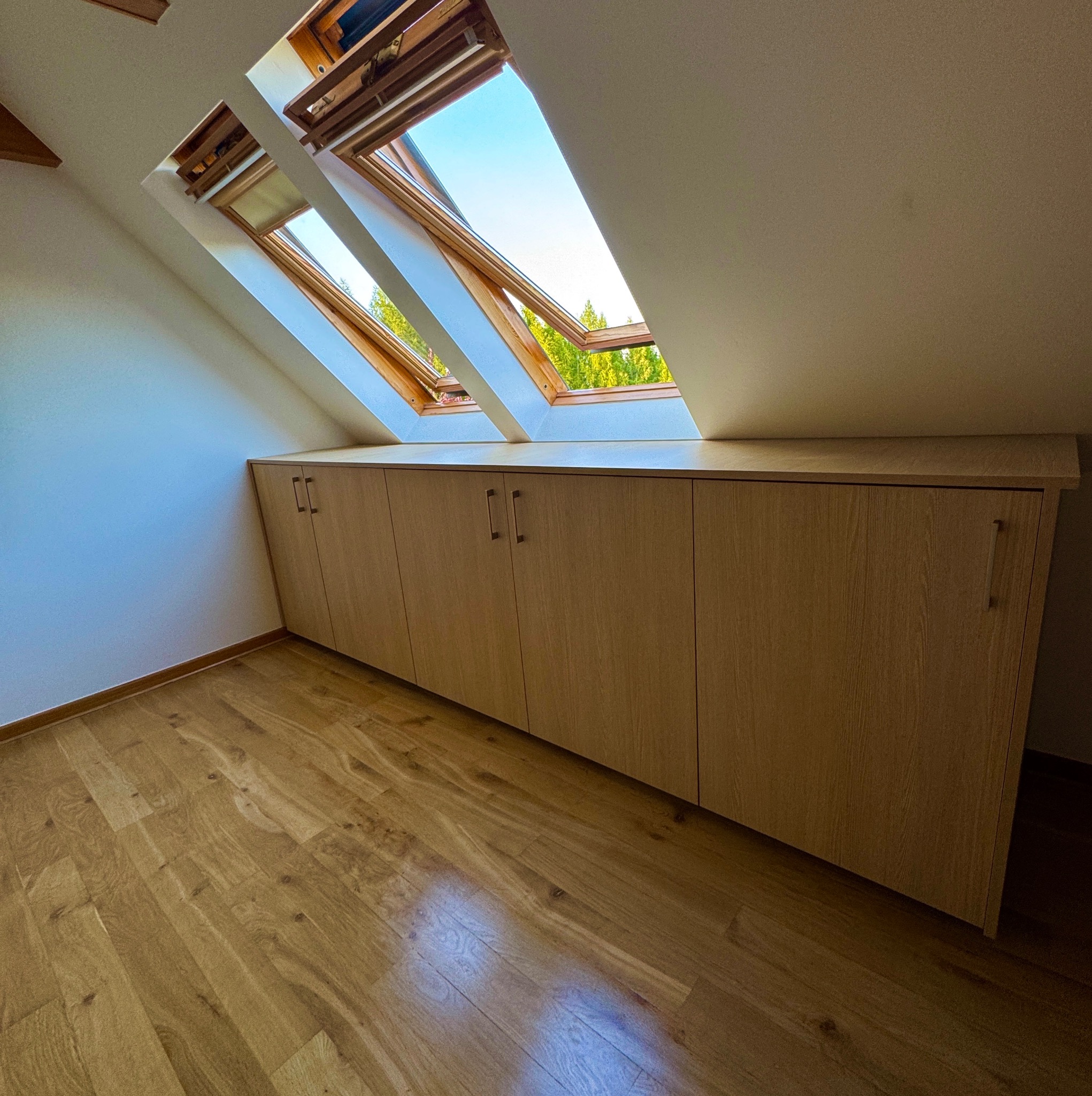 Szafa wnękowa z jasnego drewna, umieszczona pod oknami dachowymi na poddaszu. Minimalistyczne uchwyty, drewniana podłoga, skosy ścian. Widok na zieleń za oknami.