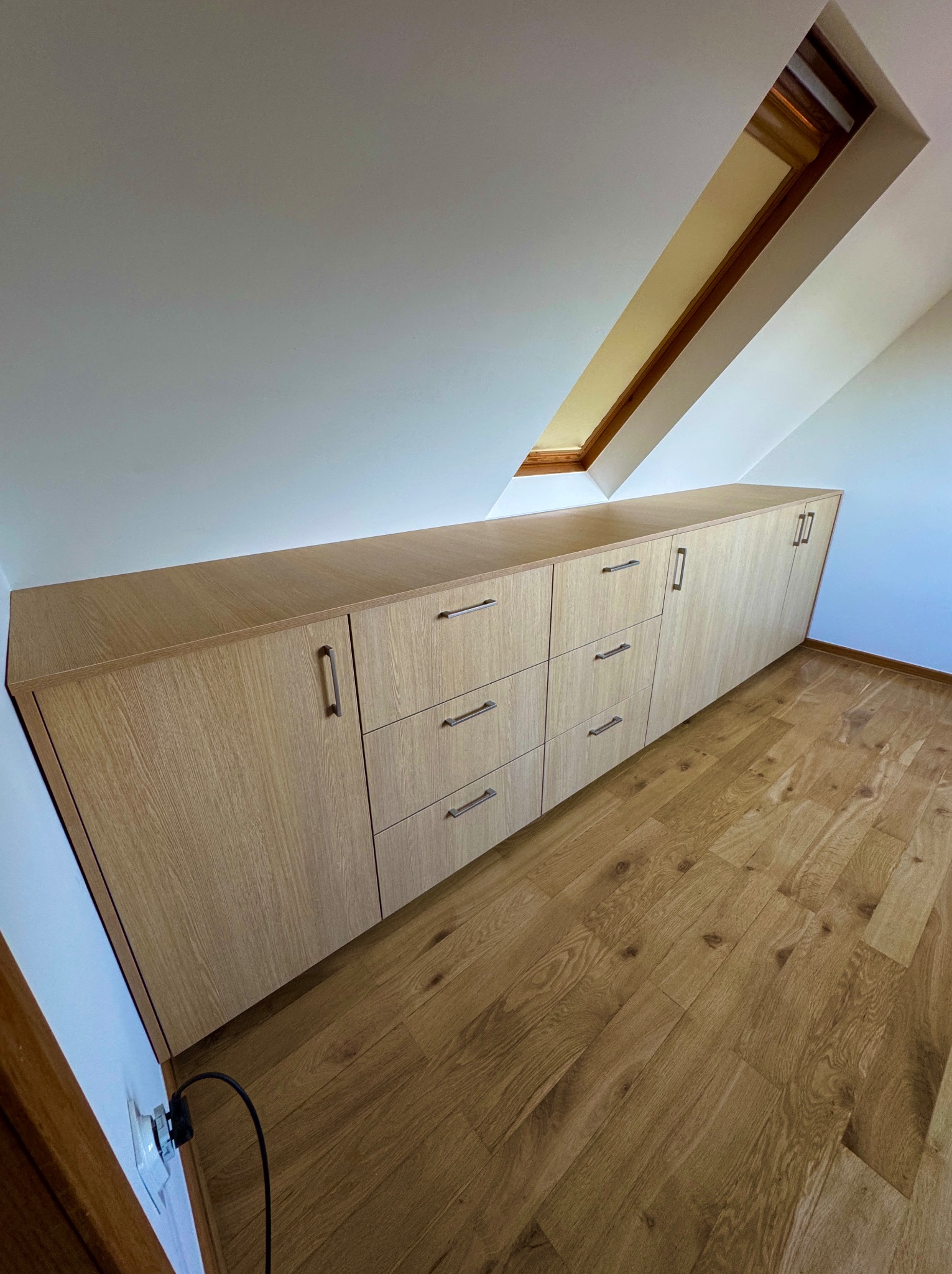 Szafa na wymiar z jasnego drewna, wkomponowana w skos dachu na poddaszu. Widoczne metalowe uchwyty, drewniana podłoga i okno dachowe.