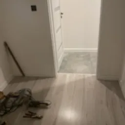 Jasne panele podłogowe w korytarzu, widoczne narzędzia monterskie i kabel w rogu. Świeżo wykończone wnętrze z białymi ścianami i drzwiami.