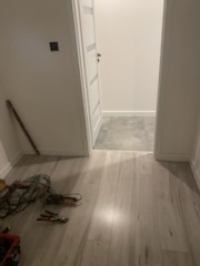 Jasne panele podłogowe w korytarzu, widoczne narzędzia monterskie i kabel w rogu. Świeżo wykończone wnętrze z białymi ścianami i drzwiami.