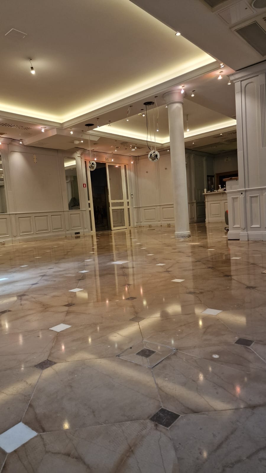 Wypolerowana marmurowa podłoga w przestronnym, jasnym pomieszczeniu z kolumnami i oświetleniem sufitowym. Widoczne odbicia światła na powierzchni podłogi.