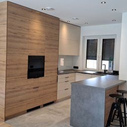 DesignLAB Manufaktura Mebli - Nowoczesna kuchnia z drewnianymi frontami szafek, czarnym piekarnikiem i szarym blatem wyspy. W tle okno z roletami i minimalistyczne oświetlenie sufitowe.