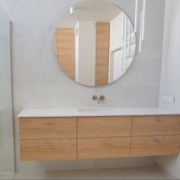 DesignLAB Manufaktura Mebli - Nowoczesna łazienka z drewnianą szafką pod umywalką i okrągłym lustrem, minimalistyczny design, białe płytki imitujące marmur, grzejnik drabinkowy.