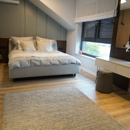 DesignLAB Manufaktura Mebli - Nowoczesna sypialnia z łazienką en-suite. Jasne drewno na podłodze i ścianach, duże okna z roletami, minimalistyczne biurko i dekoracje roślinne tworzą przytulną atmosferę.