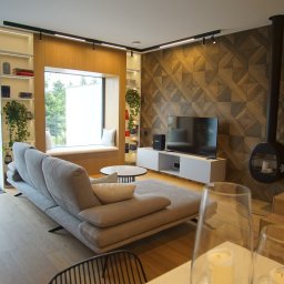 DesignLAB Manufaktura Mebli - Nowoczesny salon z szarą sofą, oknem z siedziskiem, ścianą z geometrycznym wzorem i wiszącym kominkiem. Na podłodze drewniane schody. Całość w ciepłej tonacji.