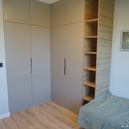 DesignLAB Manufaktura Mebli - Szara szafa narożna na wymiar z czarnymi uchwytami i drewnianym regałem w pokoju z panelami podłogowymi. Minimalistyczny design, jasne wnętrze.