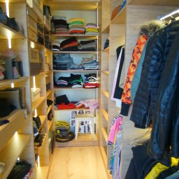 DesignLAB Manufaktura Mebli - Jasna garderoba z drewna z półkami i wieszakami, oświetlona ledami. Widoczne ubrania, buty i akcesoria. Na końcu mały stolik z ramkami.