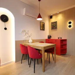 Jasne wnętrze jadalni: drewniany stół z czerwonymi krzesłami, czerwona komoda, oświetlenie punktowe i dekoracyjny zegar na ścianie z podświetleniem.