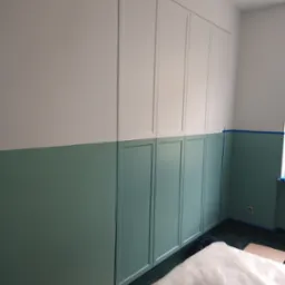 Wbudowana szafa wnękowa z panelami dekoracyjnymi, pomalowana na biało i zielono, z widocznym zabezpieczeniem podłogi i ścian taśmą malarską.