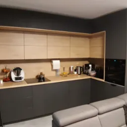 Nowoczesna kuchnia z szarymi szafkami dolnymi i drewnianymi górnymi. Widoczny sprzęt AGD i fragment szarej sofy. Ujęcie z niskiego kąta.