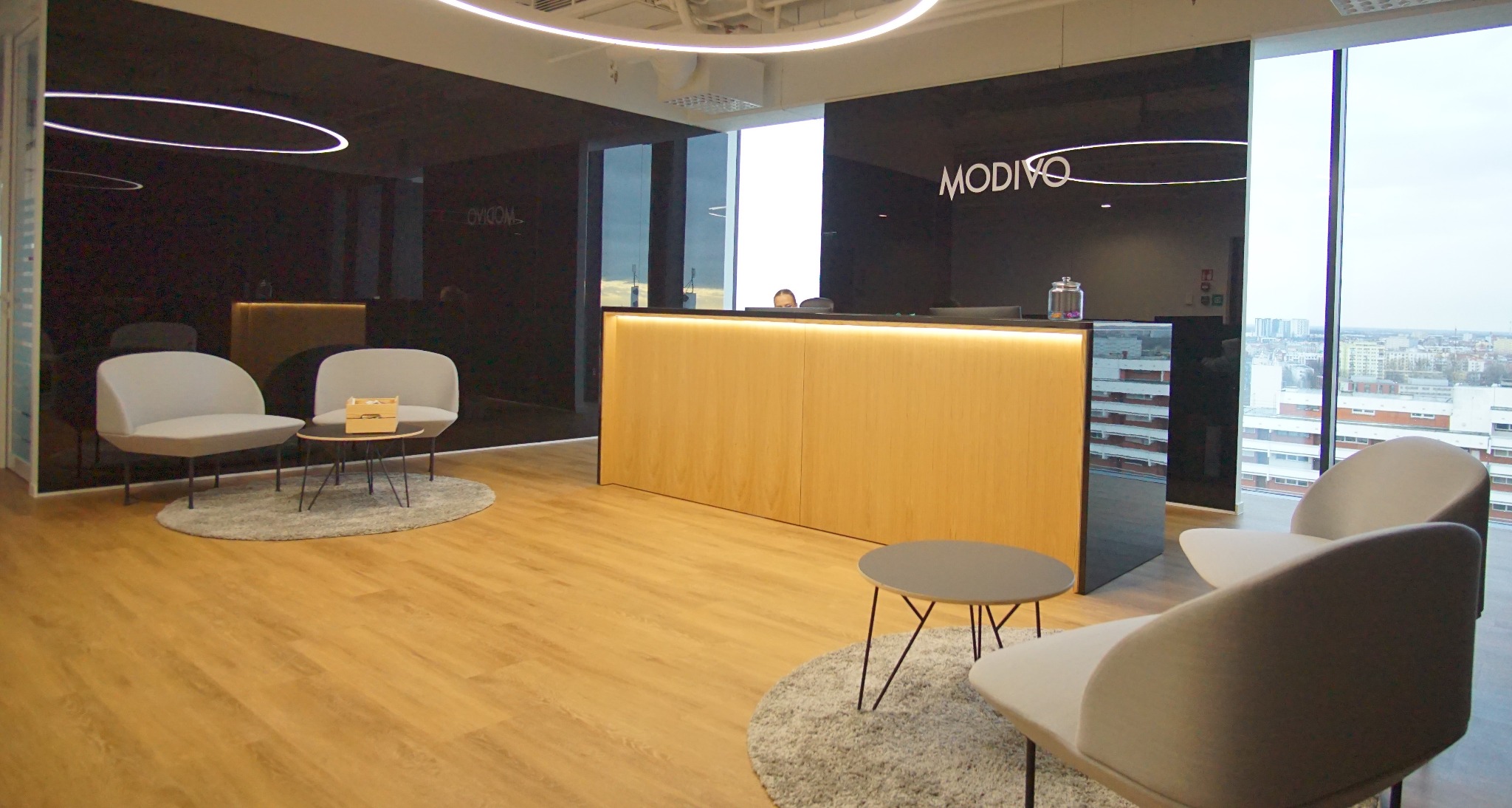 Nowoczesne wnętrze biurowe z recepcją oświetloną LED, minimalistyczne krzesła i stoliki na dywanikach, duże okna z widokiem na miasto, czarne ściany z podświetlonym logo.