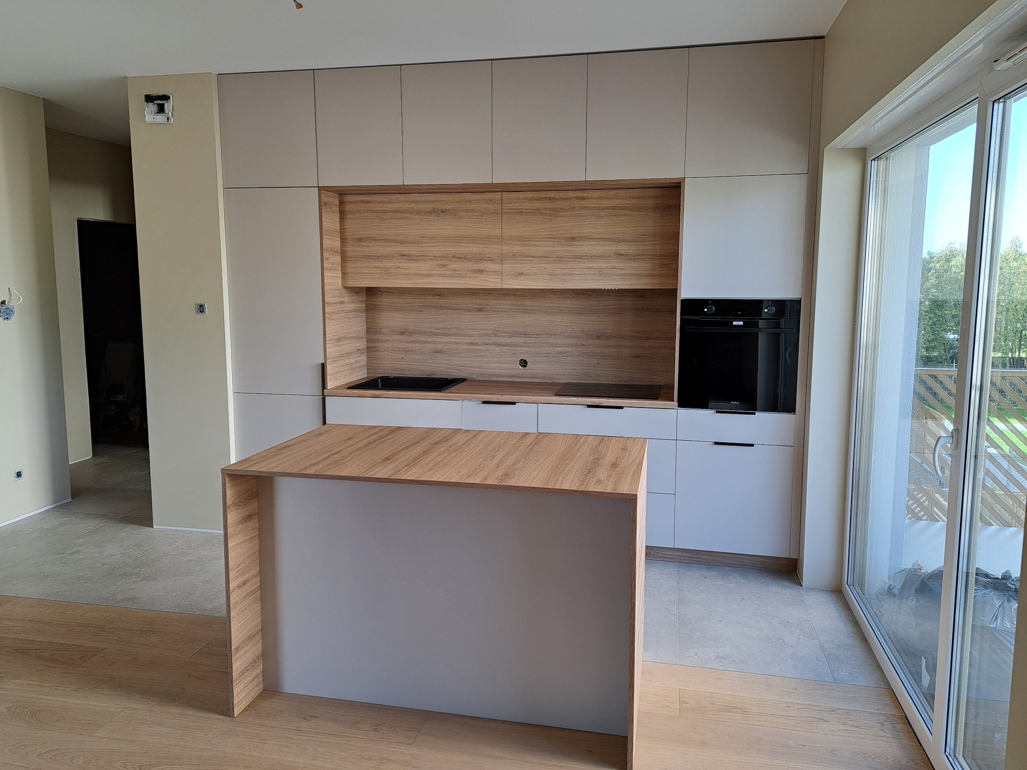 Nowoczesna kuchnia z drewnianymi akcentami. Wyspa kuchenna z blatem w kolorze drewna. Jasne szafki górne i dolne, piekarnik w zabudowie. Duże okno balkonowe.