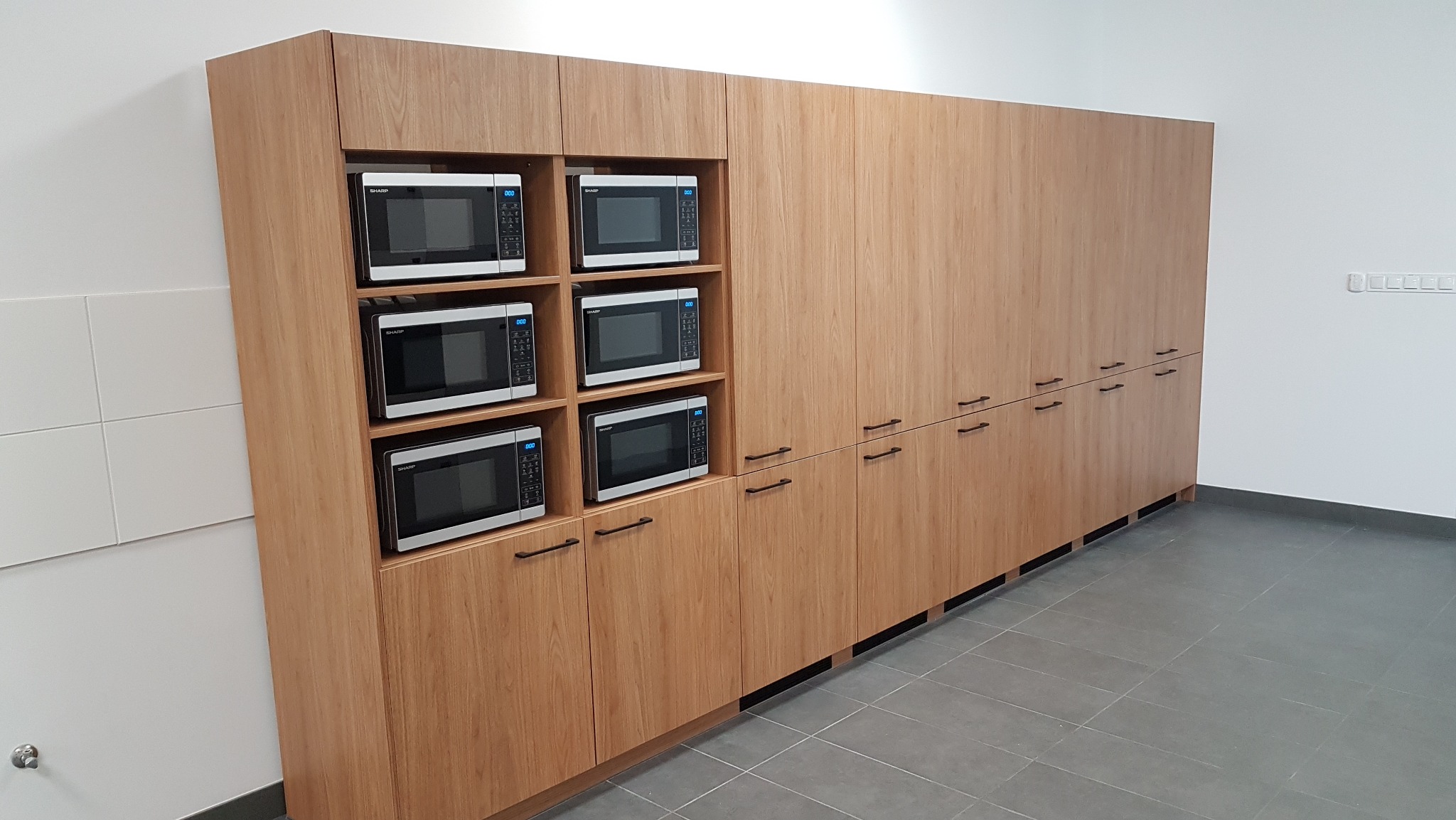 Drewniana zabudowa kuchenna z sześcioma mikrofalówkami. Minimalistyczny design, poziome uchwyty, szara podłoga i białe ściany. Naturalne światło podkreśla strukturę drewna.