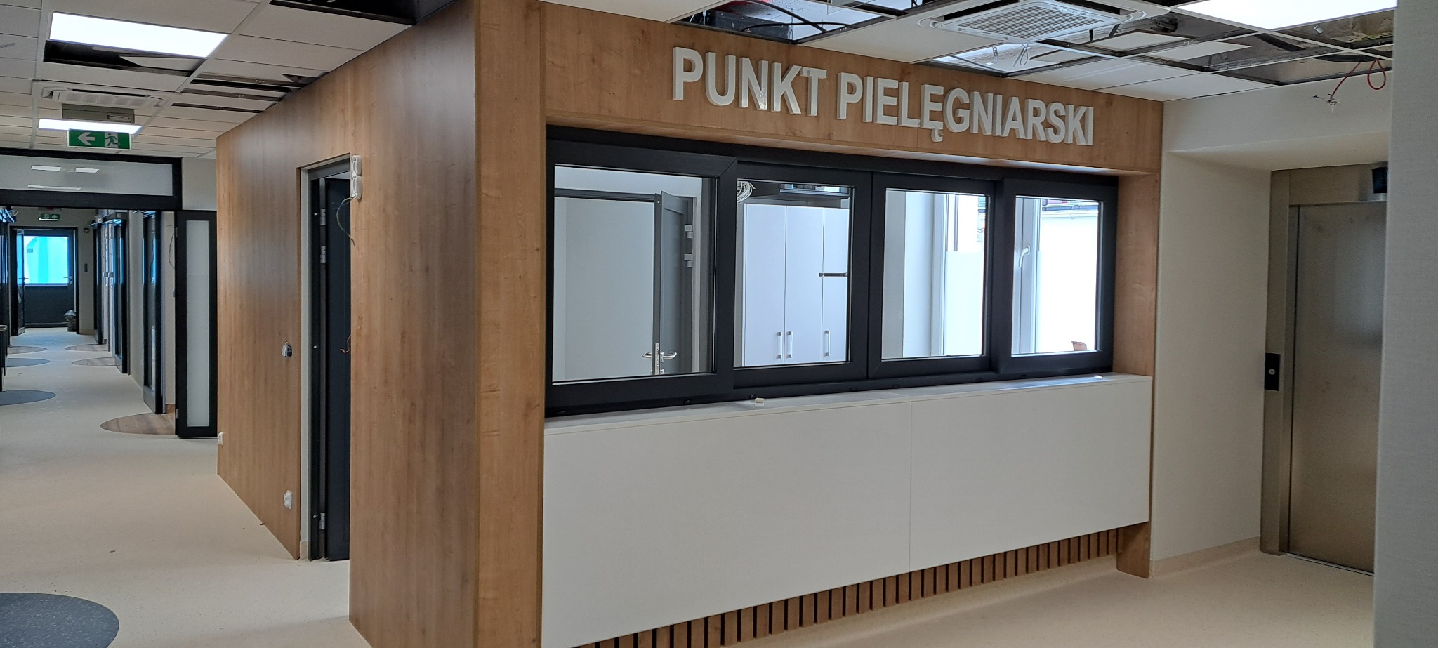 Wykończony punkt pielęgniarski z oknem i napisem, widok na korytarz szpitalny z drzwiami i windą. Drewniane i białe elementy wykończenia.