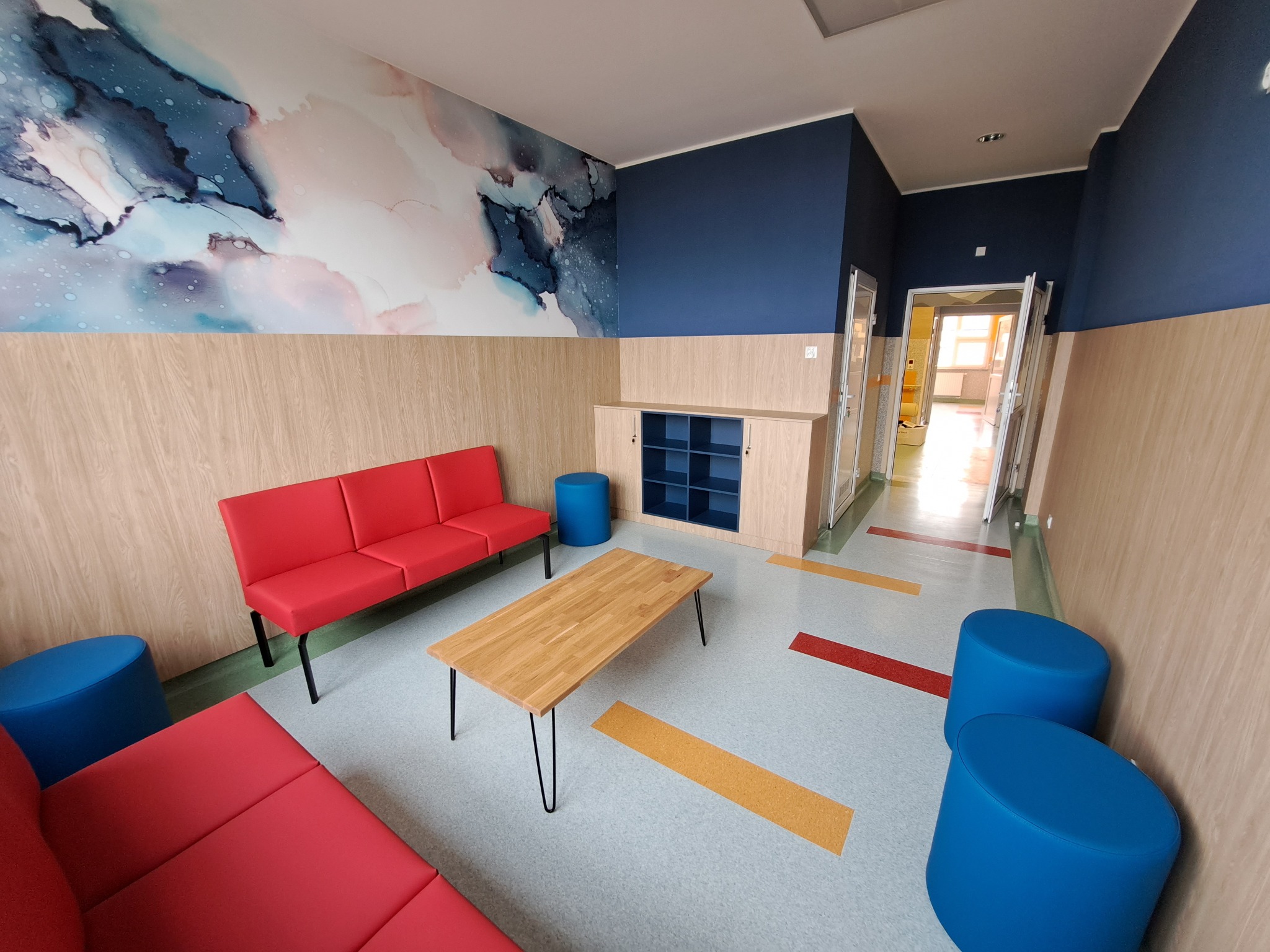 Jasne wnętrze z czerwonymi sofami, drewnianym stolikiem i niebieskimi pufami. Na ścianie abstrakcyjna grafika w odcieniach błękitu i różu, tworząca nowoczesny design.