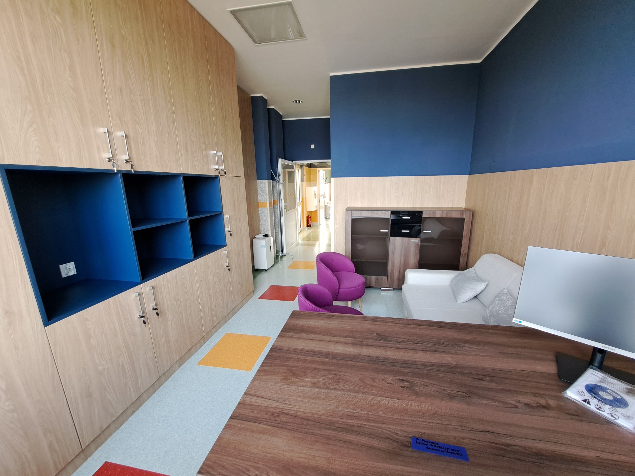 Jasne, drewniane szafy z niebieskimi wnękami, fioletowe krzesła i drewniany stół w przestronnym wnętrzu z niebieskimi akcentami na ścianach.