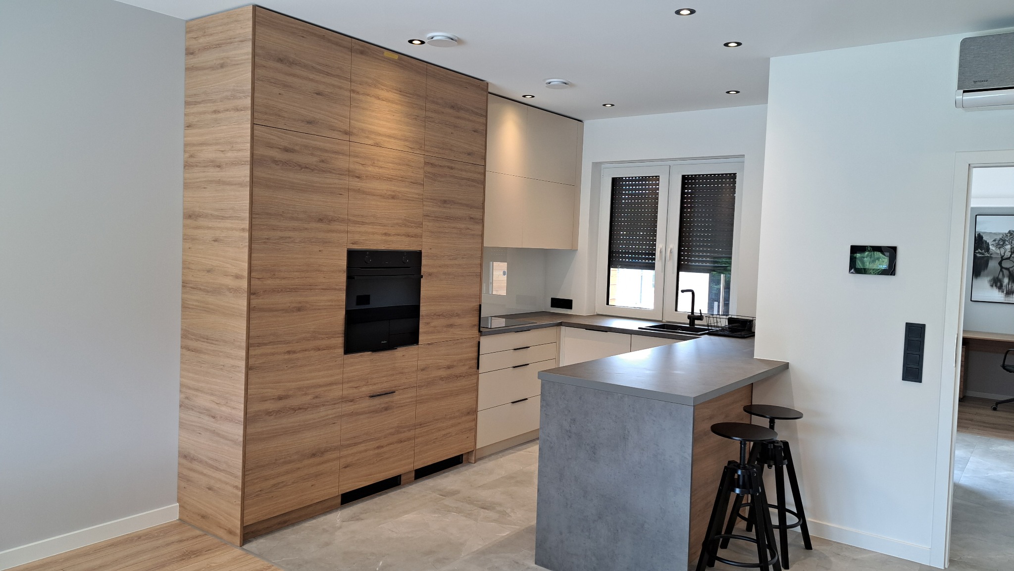 Nowoczesna kuchnia z drewnianymi frontami szafek, czarnym piekarnikiem i szarym blatem wyspy. W tle okno z roletami i minimalistyczne oświetlenie sufitowe.