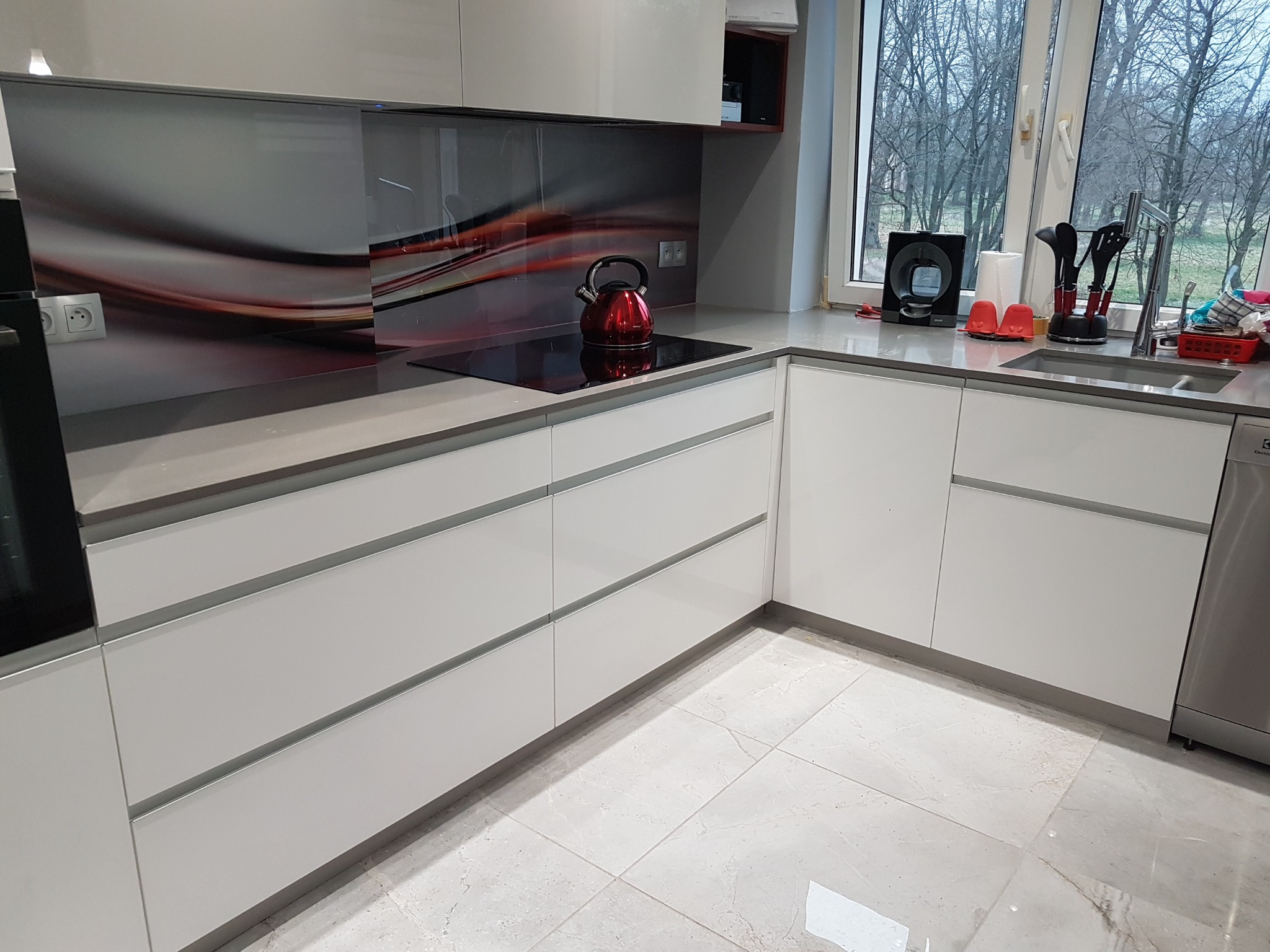 Nowoczesna kuchnia z białymi frontami, szarym blatem i abstrakcyjnym panelem nad indukcją, na której stoi czerwony czajnik. Widok na okno.